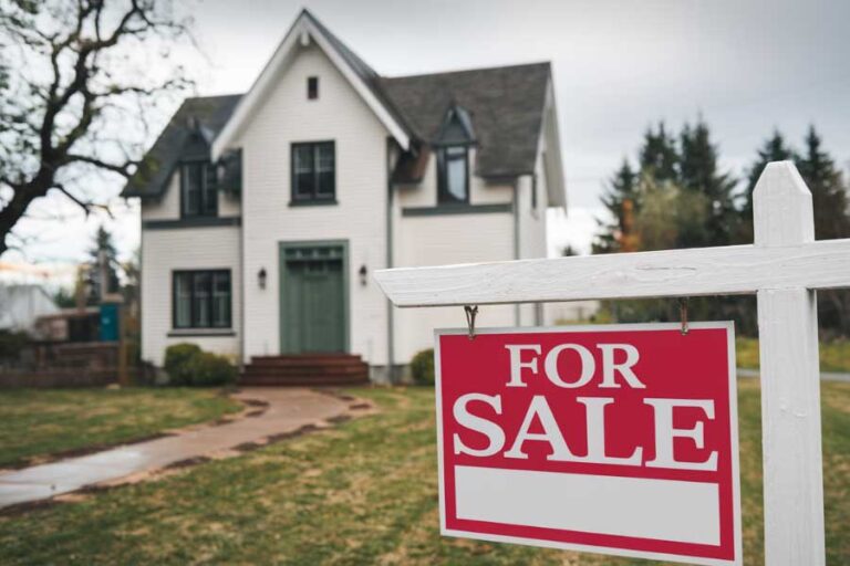 Small white house in back ground with a for sales sign in the foreground.