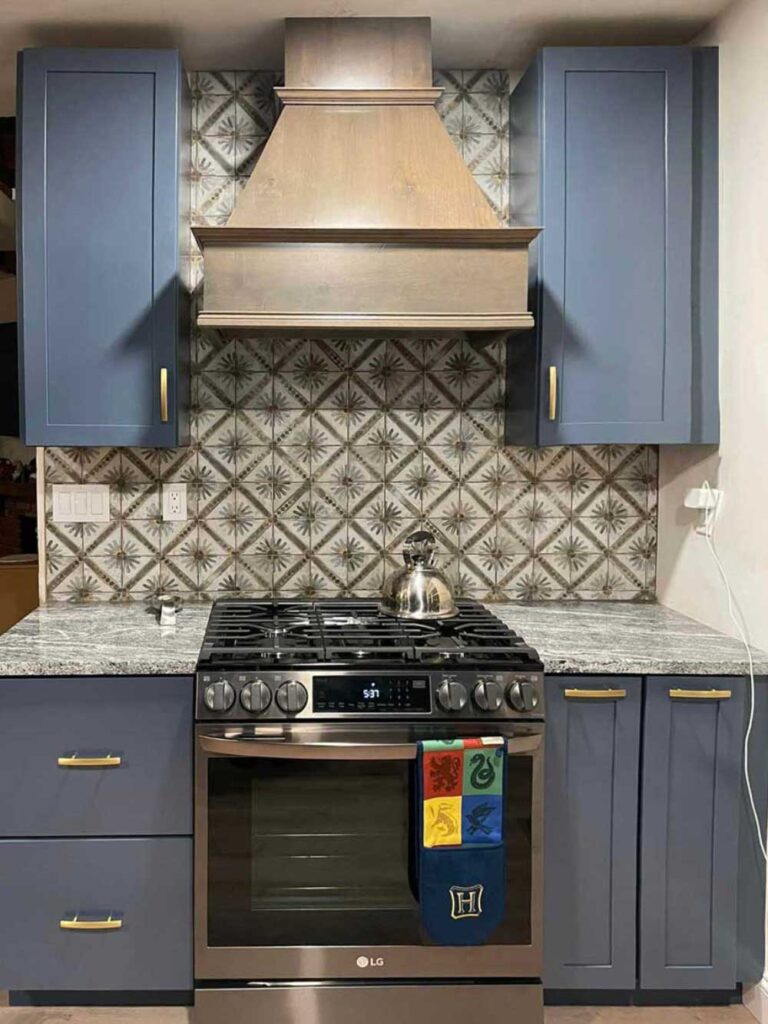 kitchen stove area with over range hood, a tiled backsplash, blue cabinets, gold hardware and granite countertops.